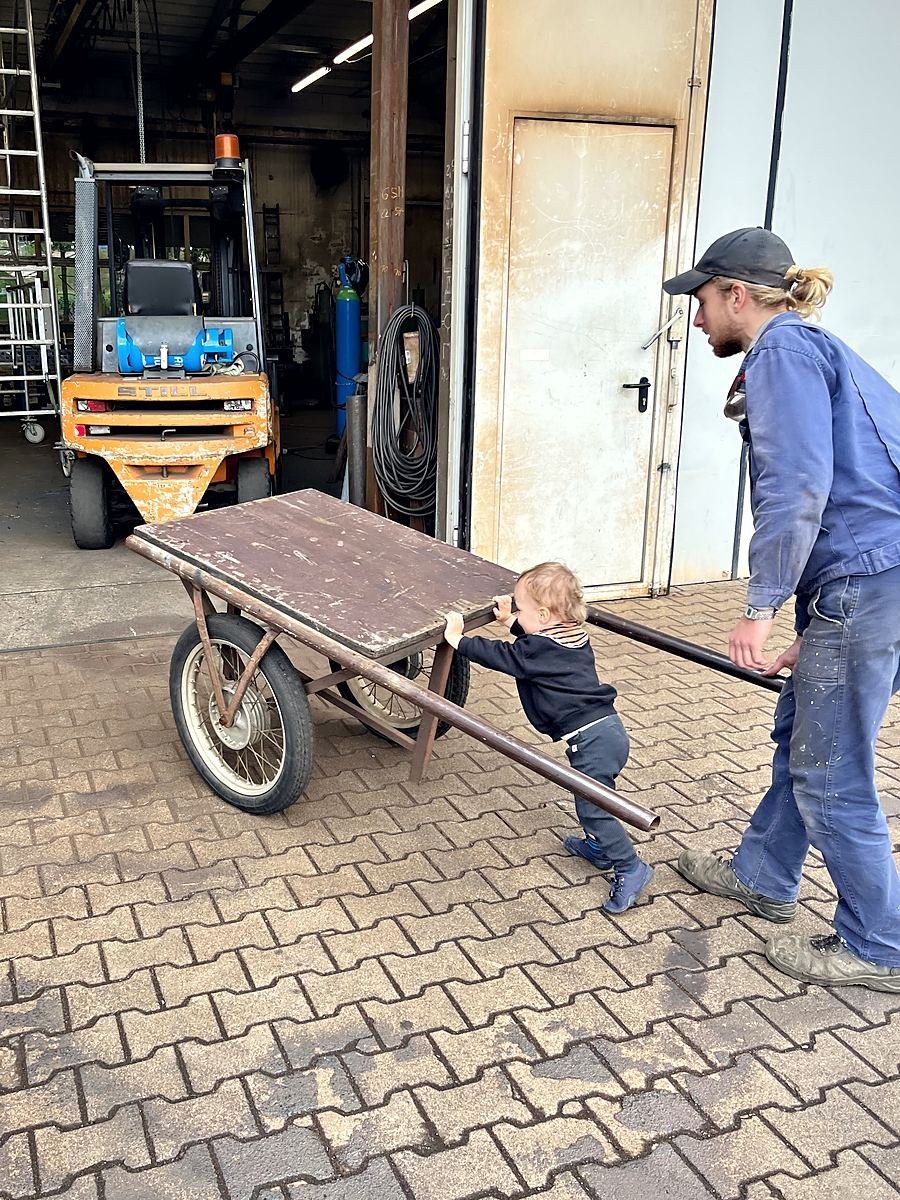 Mitarbeiter transportiert Stahlrohre, ein Kind schiebt den Handwagen mit