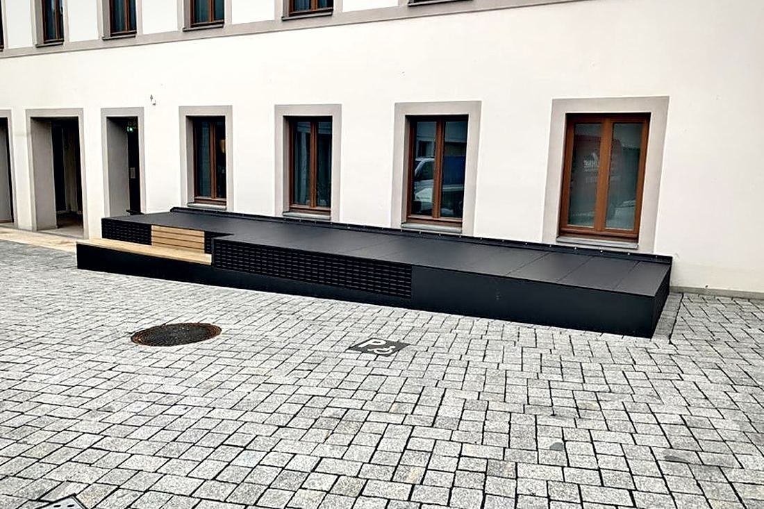 Lange Sitzanlage aus schwarzem Stahl mit Holzauflage vor einer Fassade